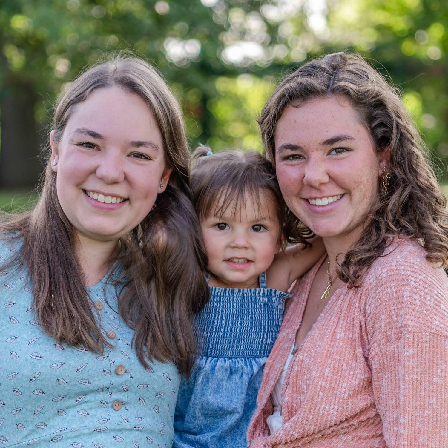 Two women and a child standing in a grassy outdoor area with trees in the background