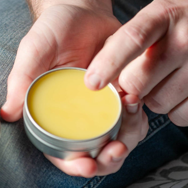 Person holding a small container  balm with a lid, sitting on a patterned surface.