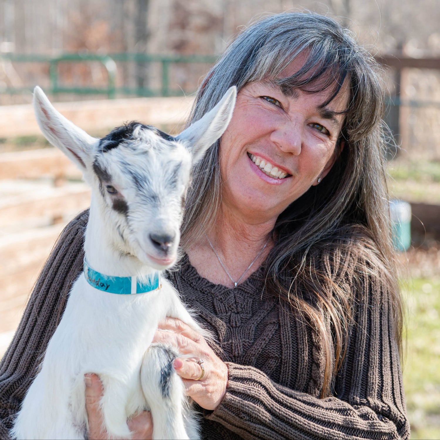 Woman holding a baby goat in an outdoor setting with trees and a fence in the background