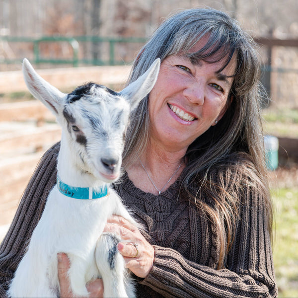 Woman holding a baby goat in an outdoor setting with trees and a fence in the background