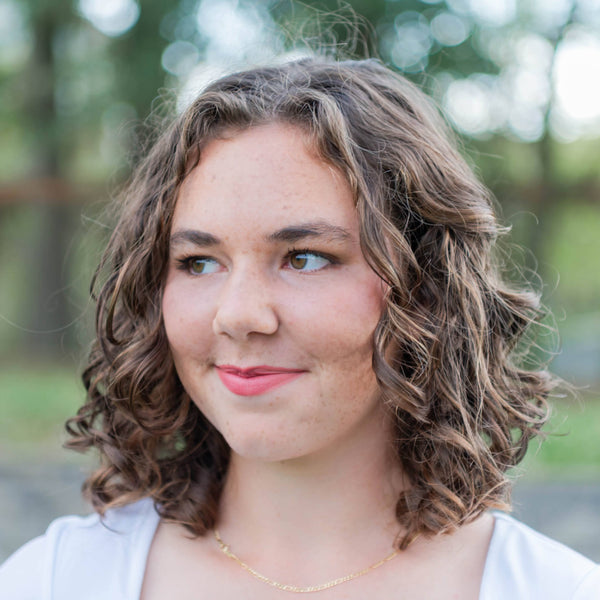 Woman with curly hair wearing a white shirt outdoors with trees in the background