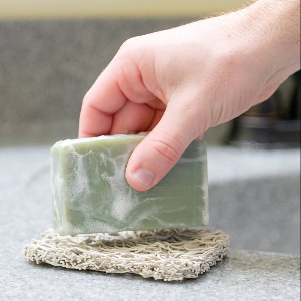 Hand holding a bar of soap over a soap rest on a kitchen counter.