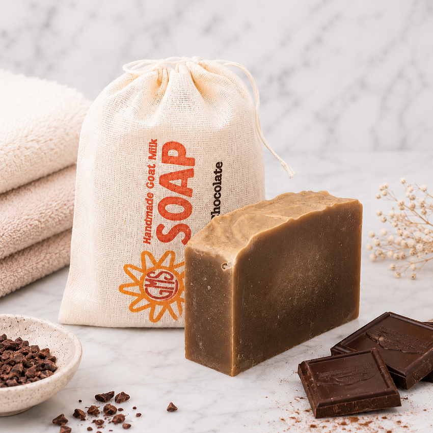 Bar of goat milk soap with a drawstring bag labeled 'Handmade Goat Milk Soap' and chocolate pieces on a marble surface.
