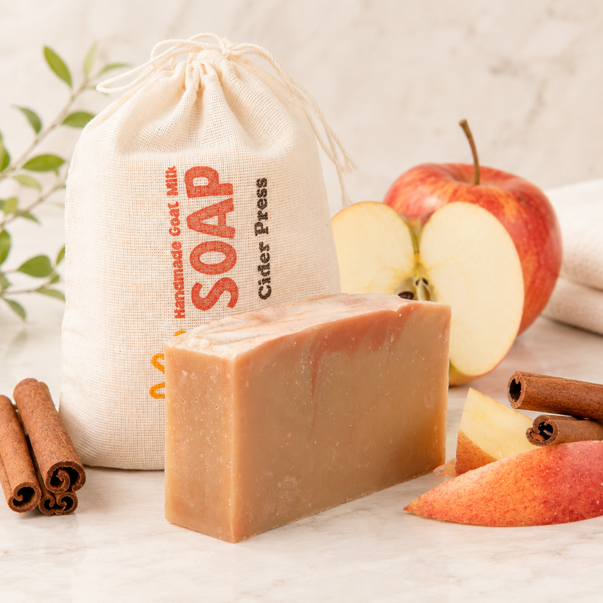 Bar of goat milk soap with an apple, cinnamon sticks, and a drawstring bag labeled 'Soap' on a light background.