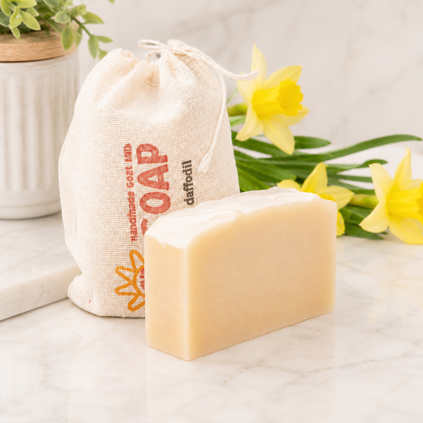 Bar of goat milk soap with a drawstring bag labeled 'Handmade Goat Milk Soap' on a light surface with yellow daffodil flowers in the background.