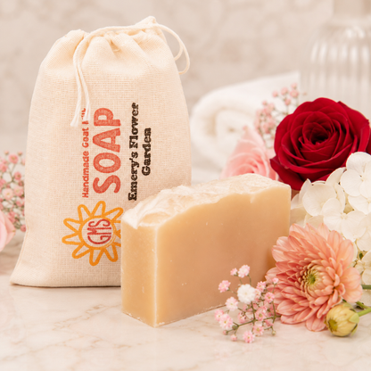 Bar of goat milk soap with a drawstring bag labeled 'Handmade Goat Soap' and flowers in the background.