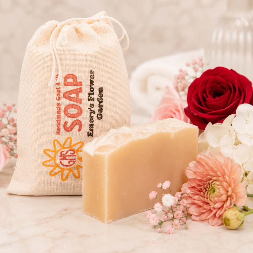 Bar of goat milk soap with a drawstring bag labeled 'Handmade Goat Soap' and flowers in the background.