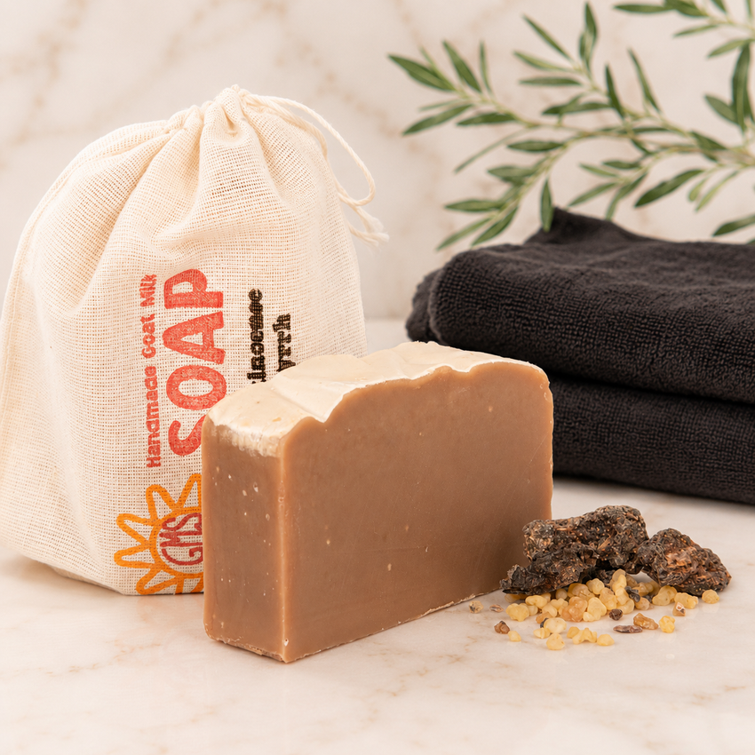 Bar of goat milk soap with a drawstring bag labeled 'Handmade Soap' and black towels in the background.