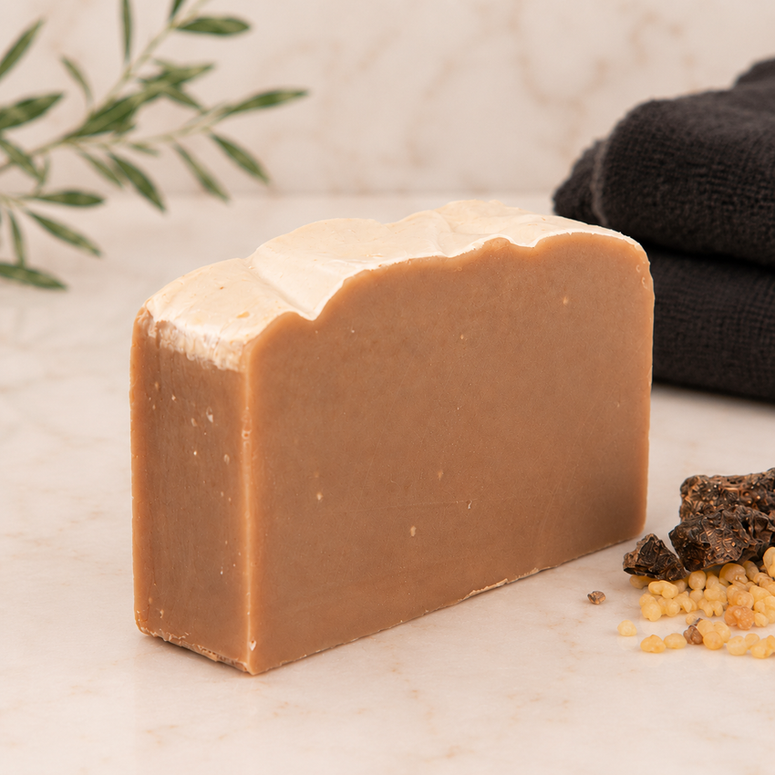 Bar of goat milk soap on a light surface with a towel and herbs in the background