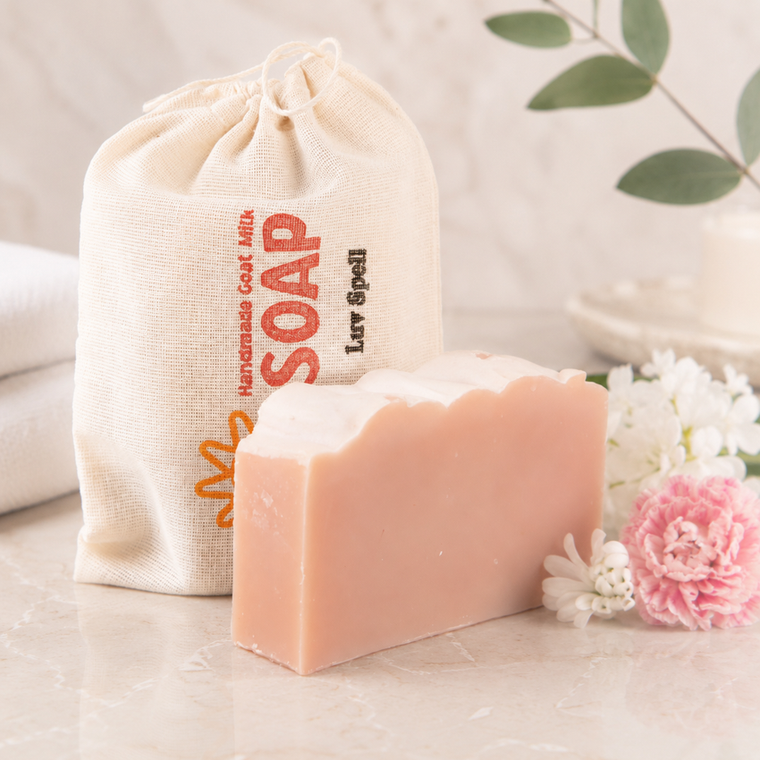 Bar of goat milk soap with a drawstring bag labeled 'SOAP' on a light surface with flowers.