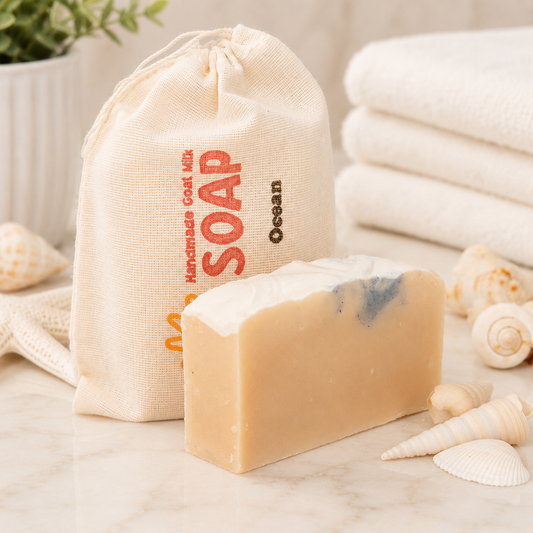 Bar of goat milk soap with a drawstring bag labeled 'Handmade Coal Milk Soap Ocean' on a surface with seashells and towels.