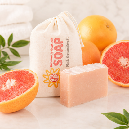 Bar of goat milk soap with a pink grapefruit theme, surrounded by pink and orange grapefruits and a drawstring bag labeled 'Pink Grapefruit Soap'.