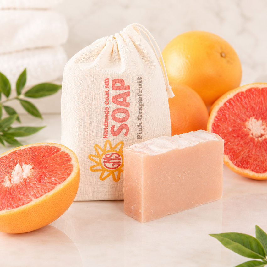 Bar of goat milk soap with a pink grapefruit theme, surrounded by pink and orange grapefruits and a drawstring bag labeled 'Pink Grapefruit Soap'.