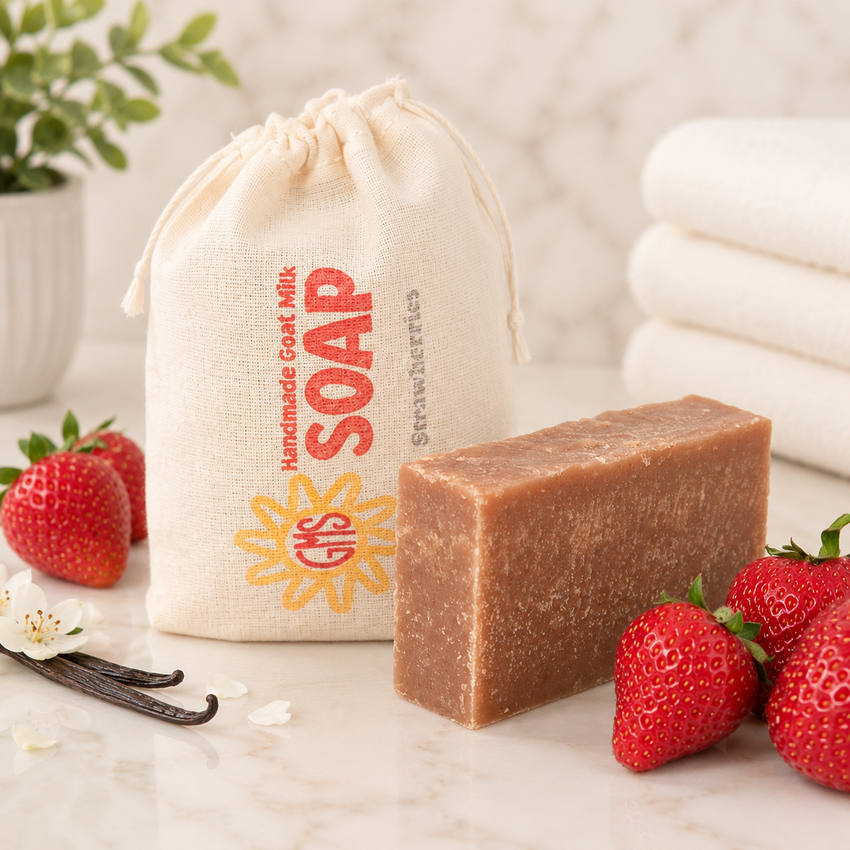Bar of goat milk soap with a drawstring bag labeled 'Handmade Goat Milk Soap' on a marble surface with strawberries and vanilla beans.