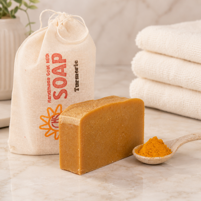 Bar of goat milk soap with a spoonful of turmeric powder on a marble surface, next to a bag labeled 'Handmade Goat Milk Soap' and folded towels.