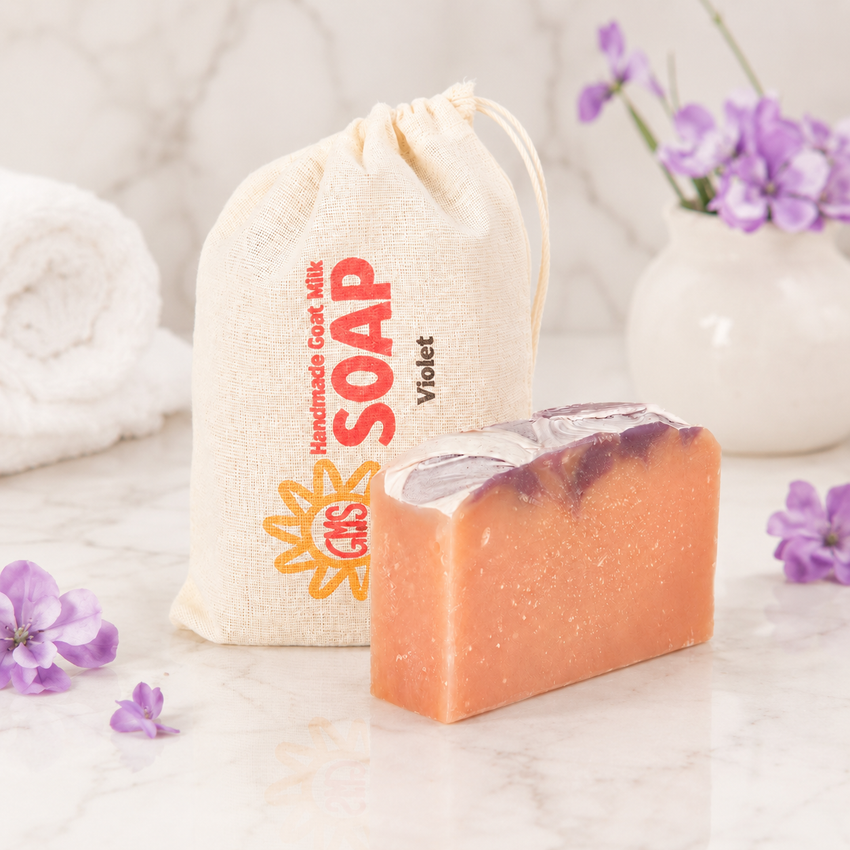 Bar of goat milk soap with a drawstring bag labeled 'Handmade Goat Milk Soap' on a light surface with Violet flowers.
