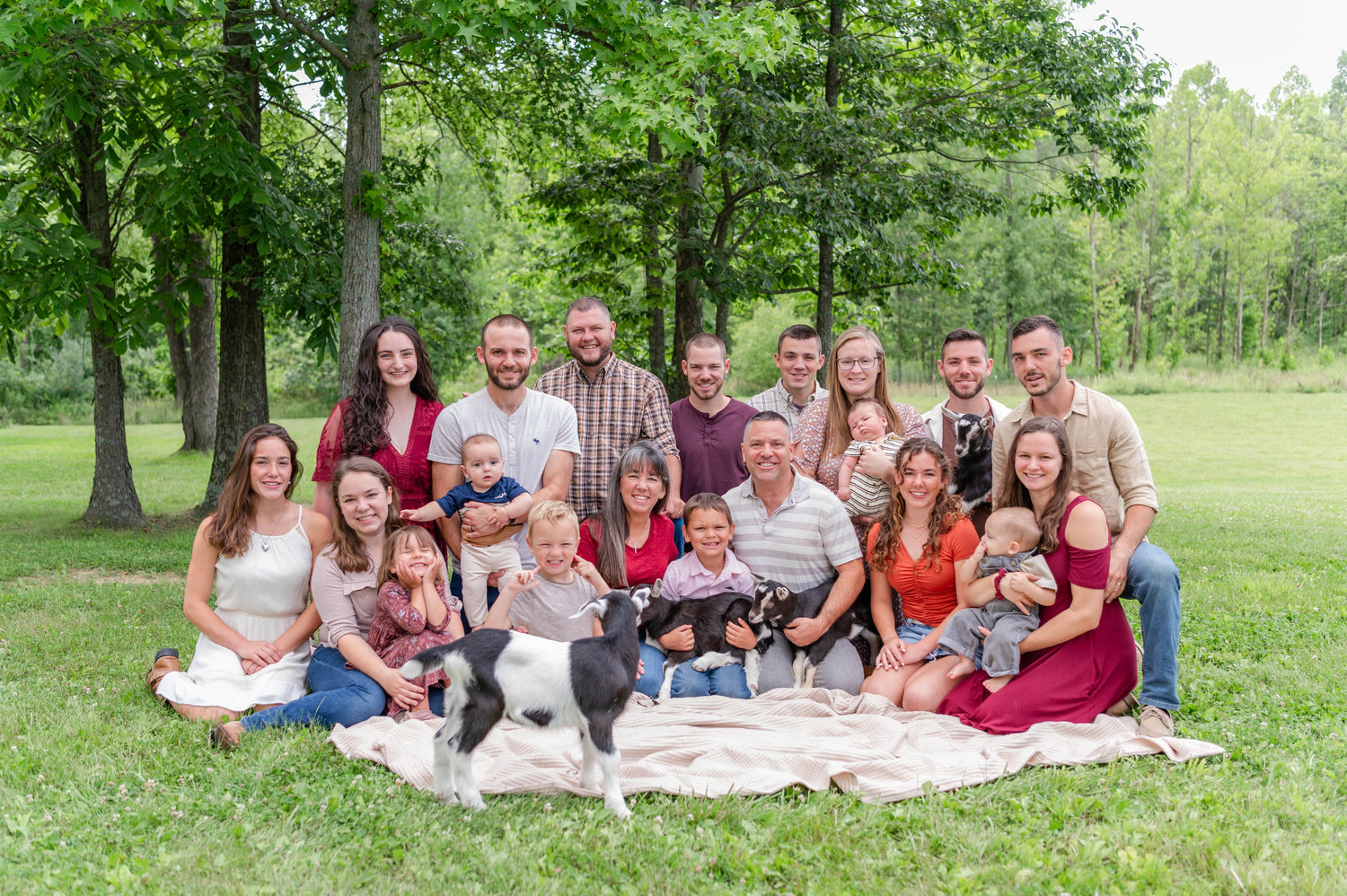 Family gathering with adults, children, and goats on a blanket in a park.