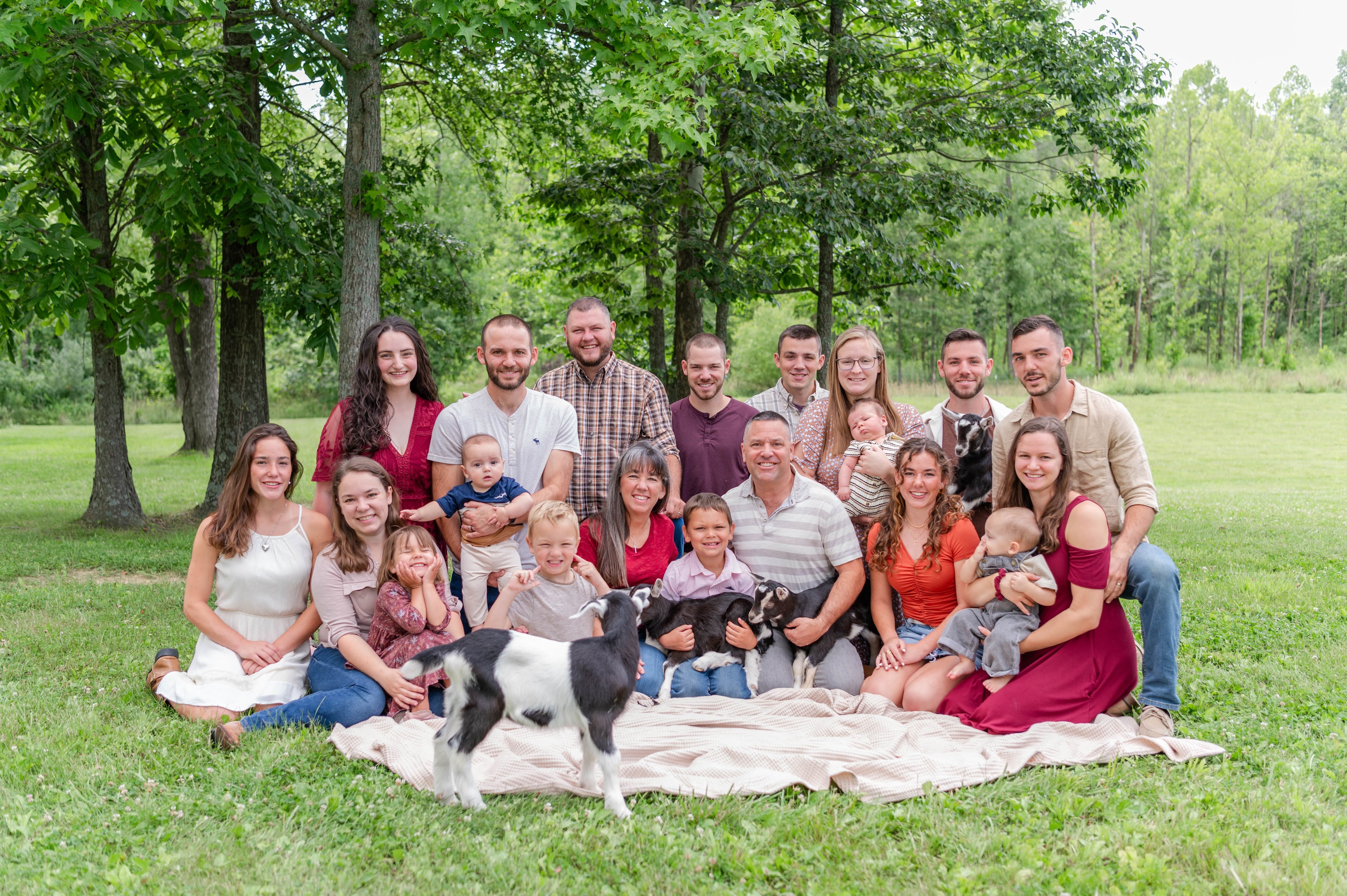 Family gathering with adults, children, and goats on a blanket in a park.